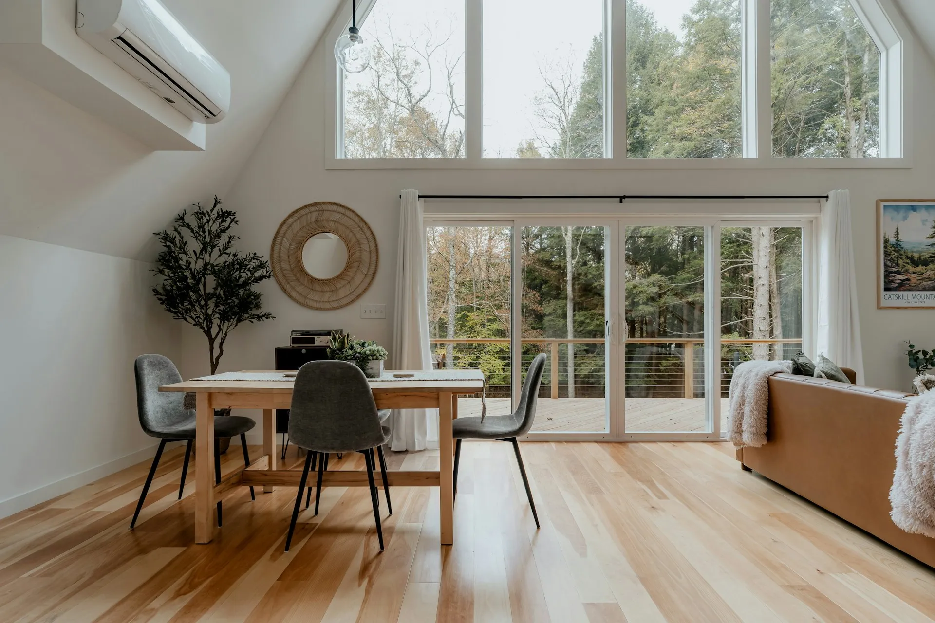 Sunlit open-plan living and dining room with pale hardwood flooring and floor-to-ceiling forest-view windows